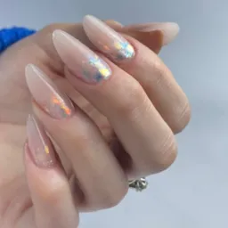 Dłoń z długimi, żelowymi paznokciami w odcieniach nude, ozdobionymi holograficznymi drobinkami, na serdecznym palcu widoczny pierścionek z oczkiem.