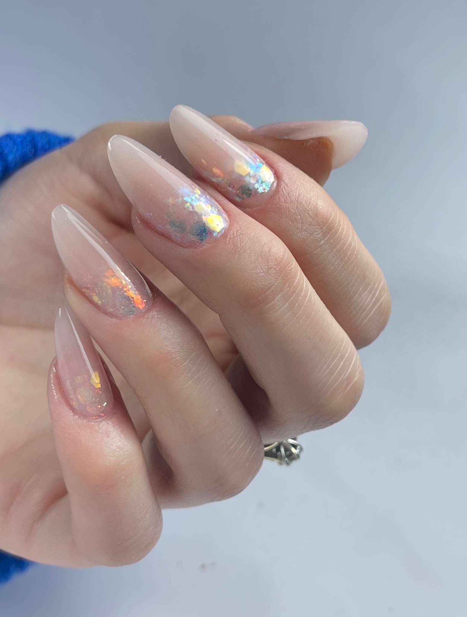 Dłoń z długimi, żelowymi paznokciami w odcieniach nude, ozdobionymi holograficznymi drobinkami, na serdecznym palcu widoczny pierścionek z oczkiem.