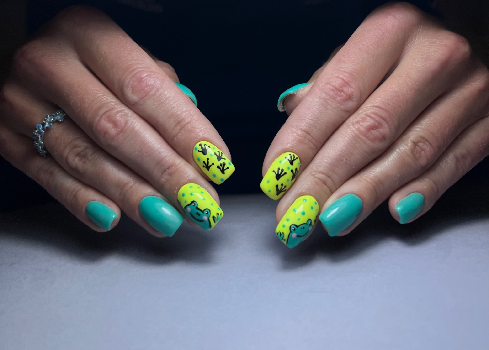 Dłonie z paznokciami pomalowanymi na jaskrawe kolory: turkusowy i neonowy żółty, z rysunkami żabek i odciskami łapek.