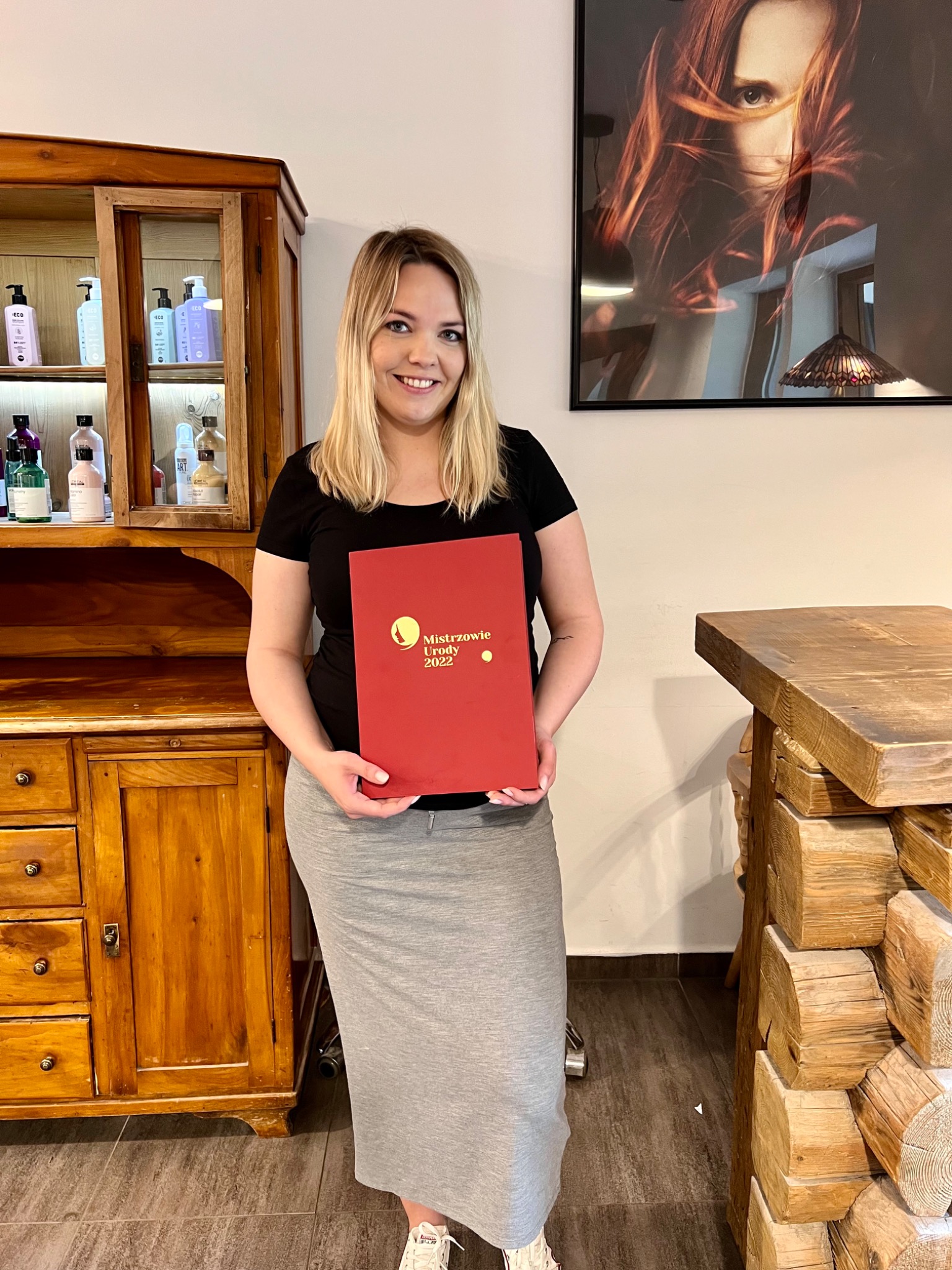 Kobieta z blond włosami trzyma w rękach czerwony dyplom 'Mistrzowie Urody 2022' na tle drewnianej komody z kosmetykami i obrazu w salonie kosmetycznym.