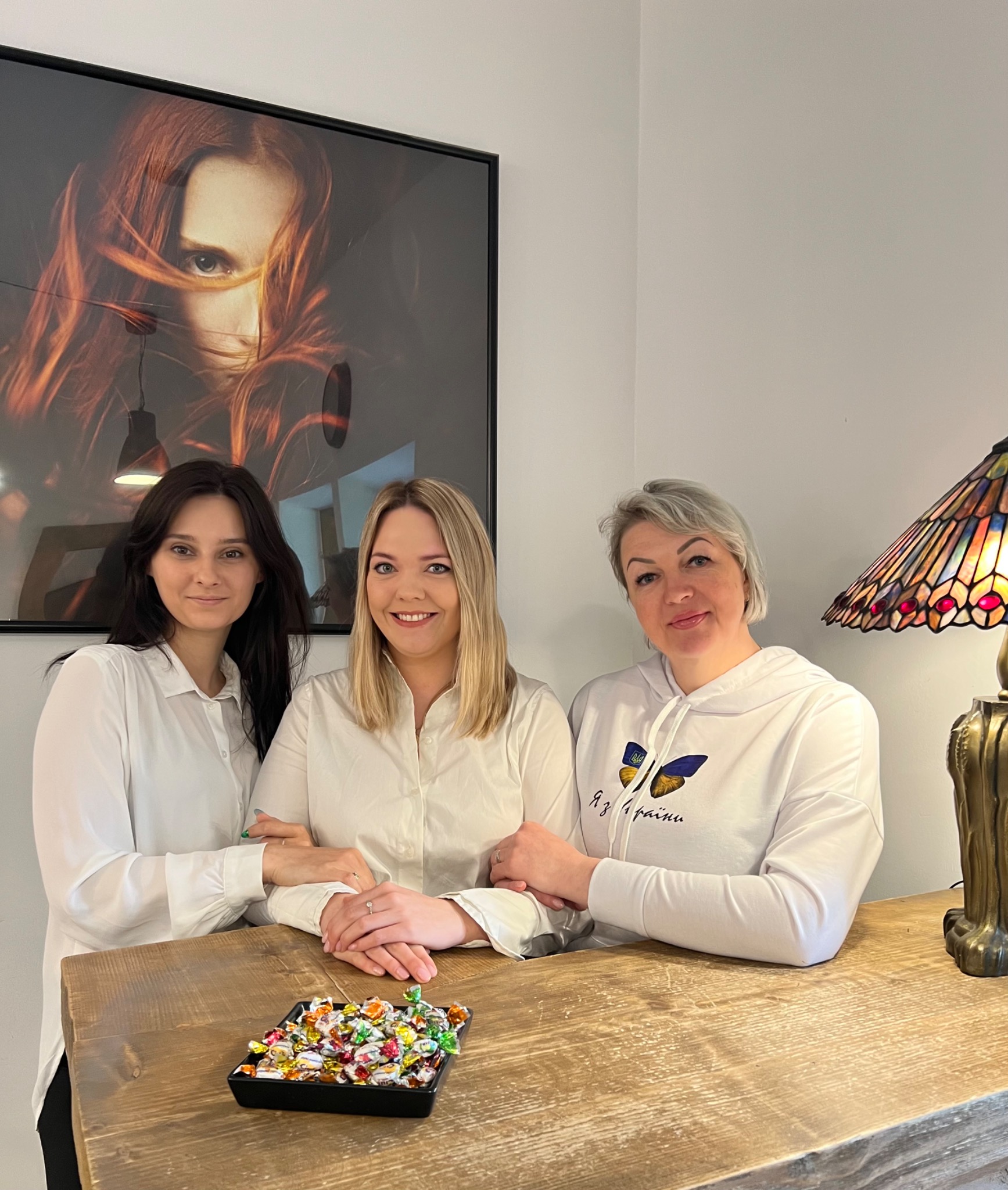 Trzy kobiety w białych koszulach stojące za drewnianym blatem z miseczką cukierków, w tle plakat z wizerunkiem kobiety z rudymi włosami, widoczna lampa witrażowa.