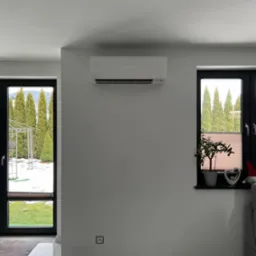 Biały klimatyzator Mitsubishi zamontowany na ścianie w jasnym pokoju z widokiem na ogród przez okno i drzwi balkonowe.