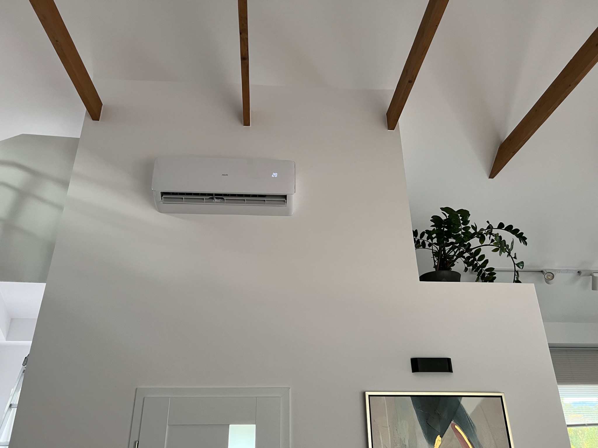 Biały klimatyzator ścienny z wyświetlaczem temperatury (28 stopni Celsjusza) zamontowany w nowoczesnym salonie z drewnianymi belkami stropowymi i rośliną doniczkową na półce.
