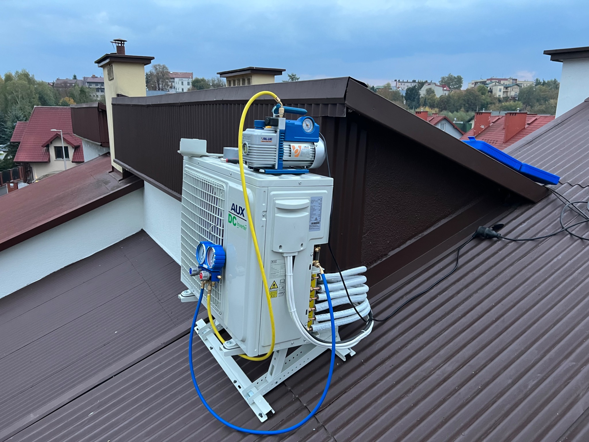 Montaż klimatyzatora AUX na brązowym dachu domu z użyciem pompy próżniowej VALUE i manometrów, widoczne przewody i instalacja chłodnicza.