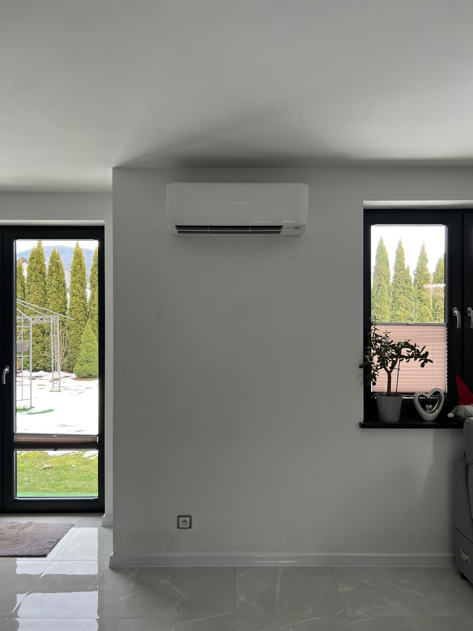 Biały klimatyzator Mitsubishi zamontowany na ścianie w jasnym pokoju z widokiem na ogród przez okno i drzwi balkonowe.