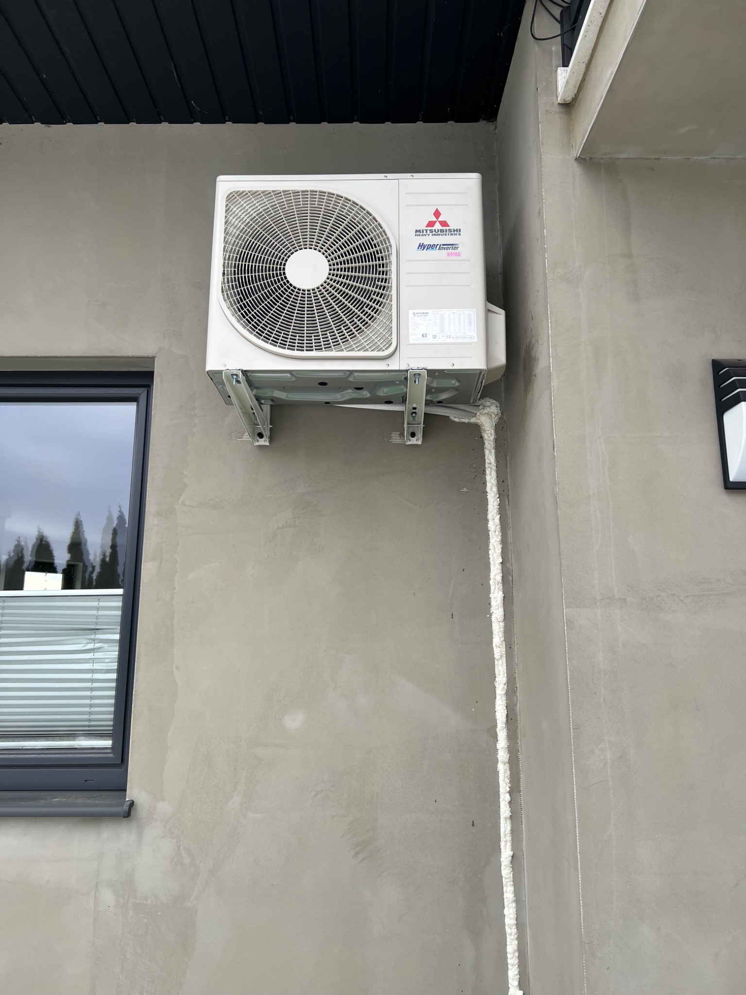 Zewnętrzna jednostka klimatyzacji Mitsubishi Hyper Inverter, zamontowana na szarej elewacji budynku, z widoczną pianką montażową uszczelniającą przewody.