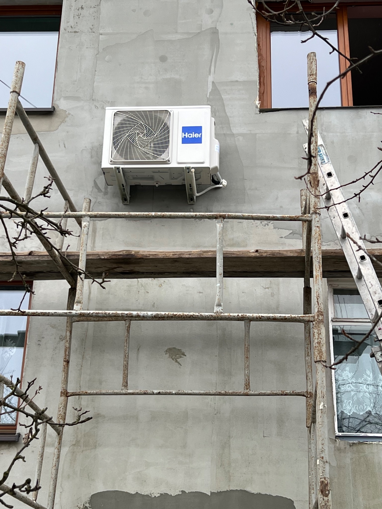 Zewnętrzna jednostka klimatyzacji Haier zamontowana na szarej ścianie budynku, widoczna z poziomu rusztowania, obok okna z brązową ramą.