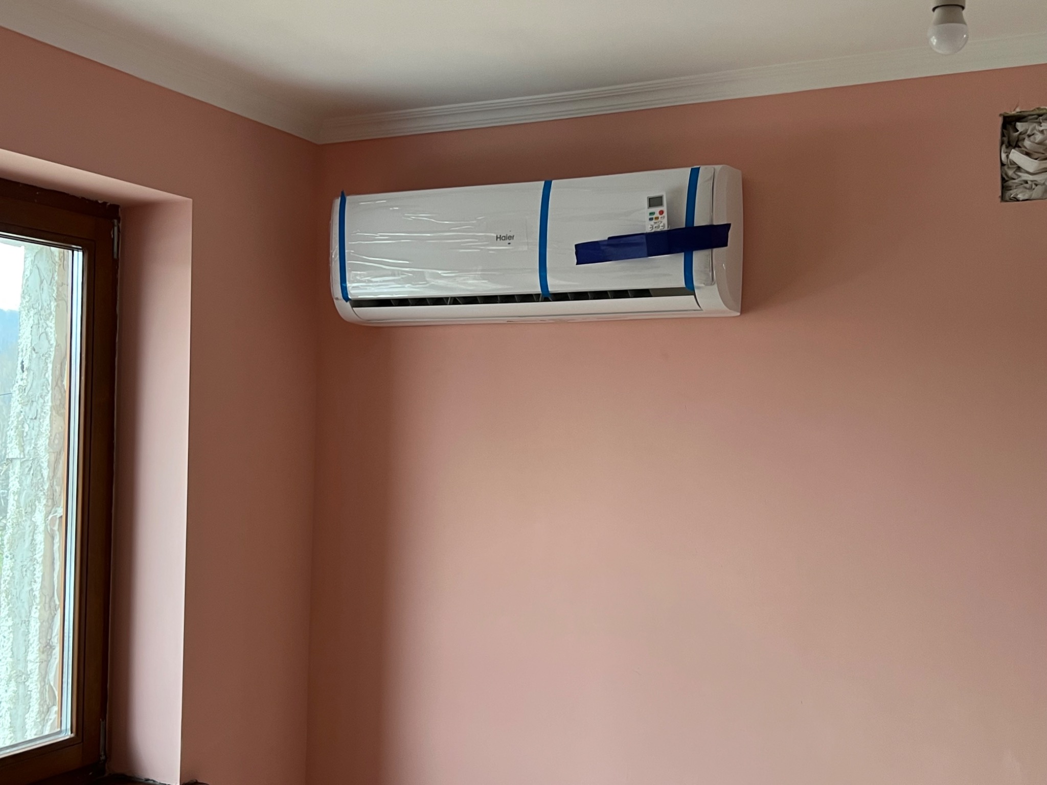 Nowy, biały klimatyzator Haier, z niebieską taśmą zabezpieczającą i folią ochronną, zamontowany na różowej ścianie w pokoju z oknem i widocznym otworem w ścianie.
