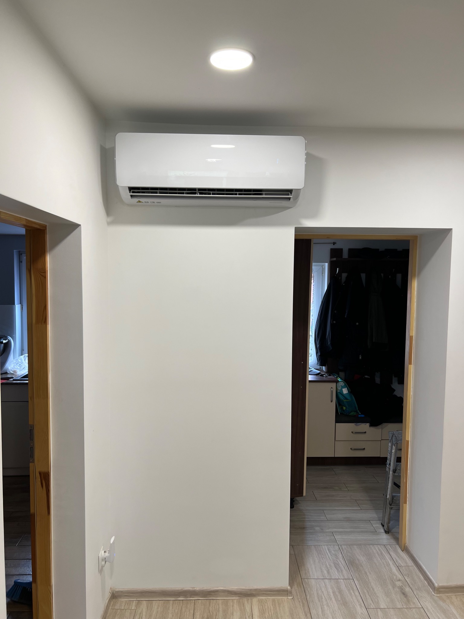 Biały klimatyzator ścienny zamontowany w przedpokoju domu, widoczny z perspektywy podłogi, z widokiem na otwarte drzwi do kuchni i garderoby.
