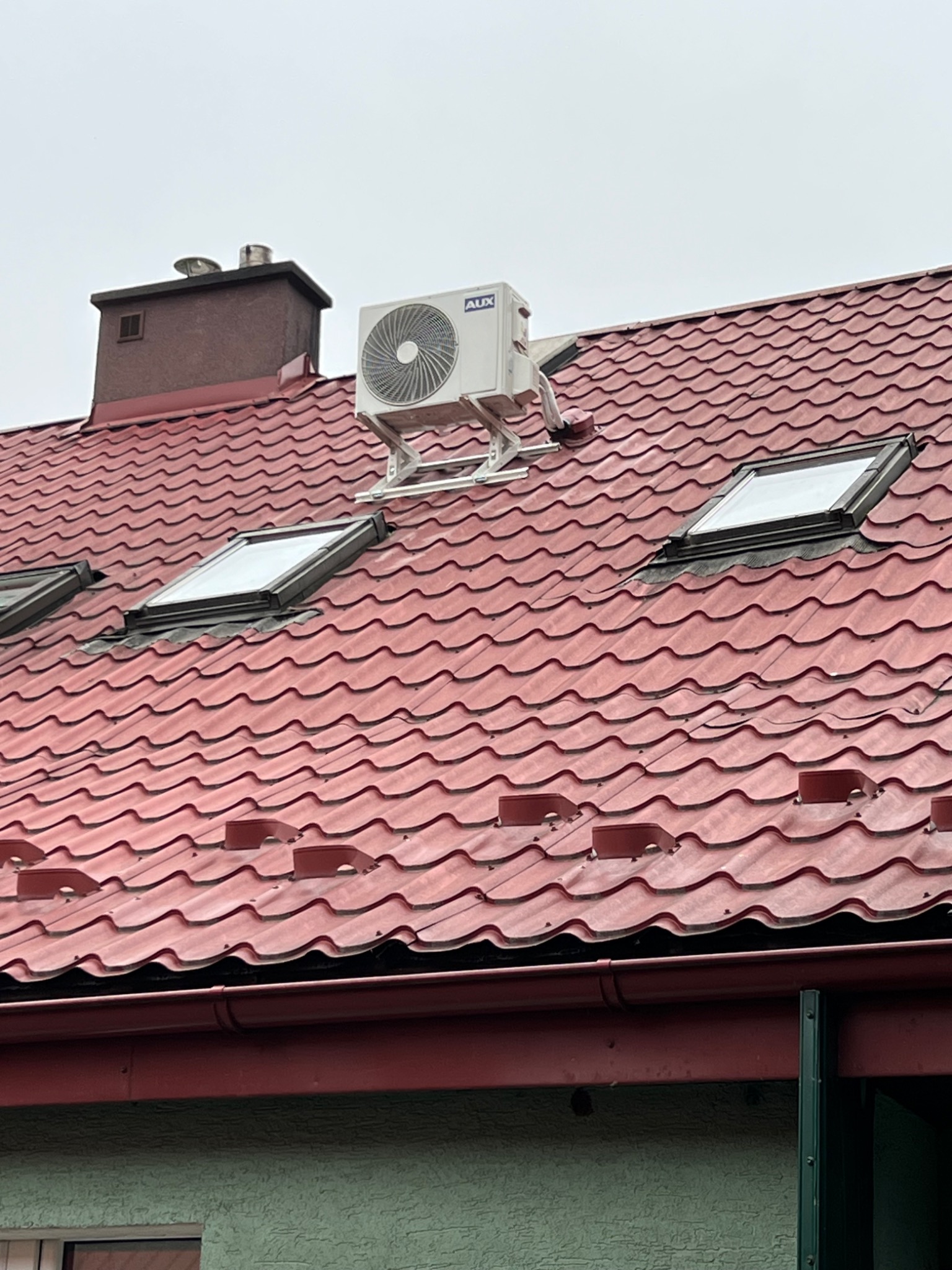 Biała jednostka zewnętrzna klimatyzatora marki AUX zamontowana na metalowym stelażu na czerwonym dachu skośnym z oknami dachowymi i kominem. Pochmurne niebo w tle.