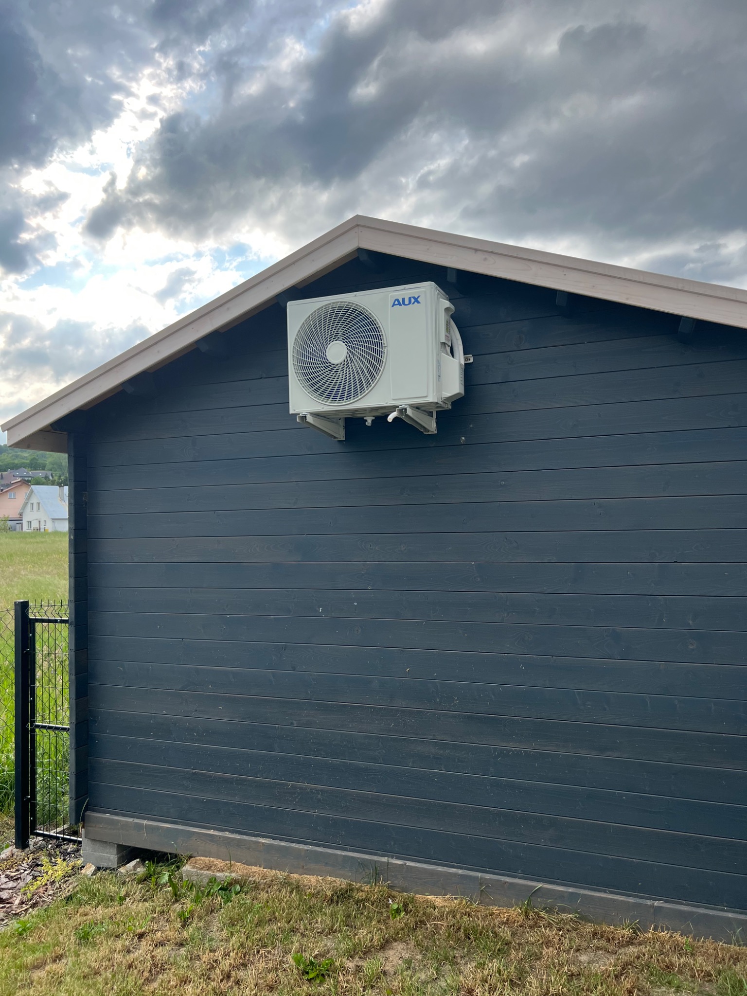 Zewnętrzna jednostka klimatyzacji marki AUX zamontowana na ciemnoszarym, drewnianym domku narzędziowym z widoczną konstrukcją dachu i fragmentem ogrodzenia.