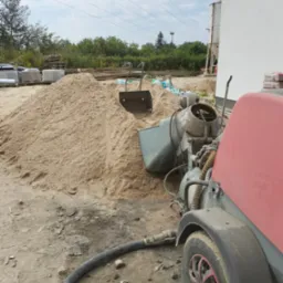 Stos piasku budowlanego obok maszyny do mieszania zaprawy z widocznym wężem, na placu budowy, w tle palety z materiałami budowlanymi i zieleń.