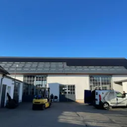 Instalacja paneli fotowoltaicznych na dachu budynku przemysłowego, widoczny wózek widłowy i furgonetka z logo firmy, pracownik na drabinie.