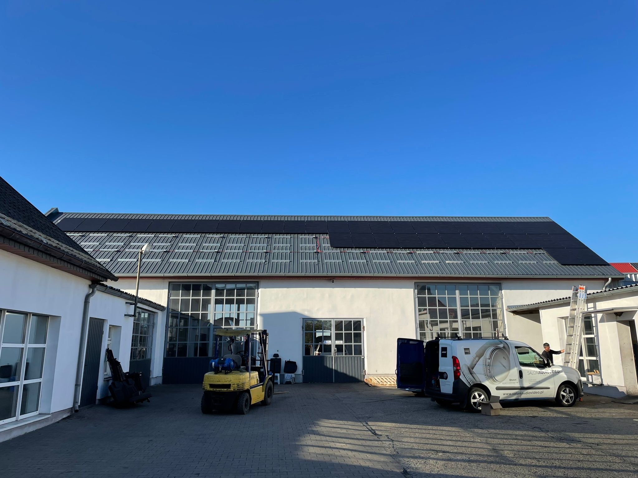 Instalacja paneli fotowoltaicznych na dachu budynku przemysłowego, widoczny wózek widłowy i furgonetka z logo firmy, pracownik na drabinie.