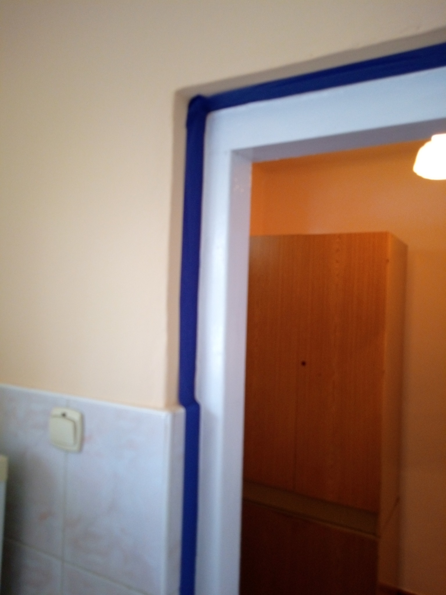 Przygotowana do malowania futryna drzwiowa, zabezpieczona niebieską taśmą malarską, widoczny fragment pomalowanej ściany w odcieniu brzoskwiniowym i fragment szafy w tle.