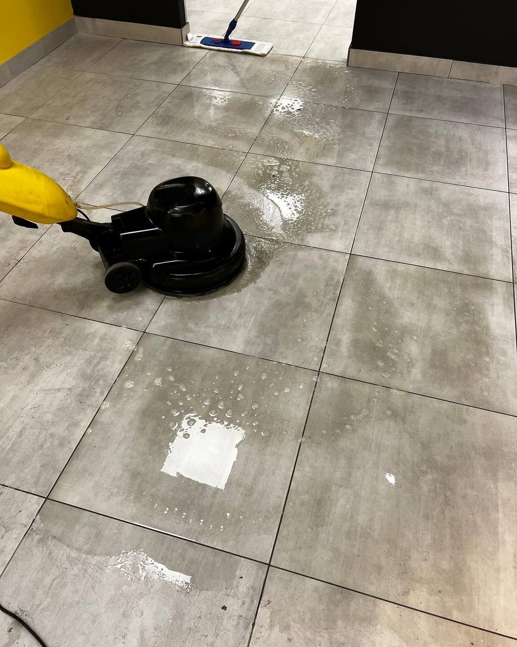 Szorowarka do podłóg w trakcie pracy na szarej, kwadratowej podłodze z płytek, widoczny mokry ślad po czyszczeniu i mop w tle.