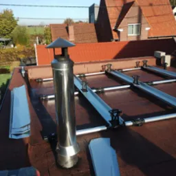 Montaż konstrukcji wsporczej pod panele fotowoltaiczne na dachu pokrytym papą, widoczny komin z nierdzewnej stali i elementy mocujące szyny.