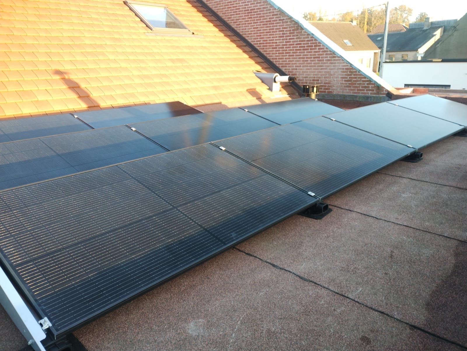 Panele fotowoltaiczne zamontowane na dachu pokrytym czerwoną dachówką, widoczne mocowania paneli i fragmenty innych budynków w tle.