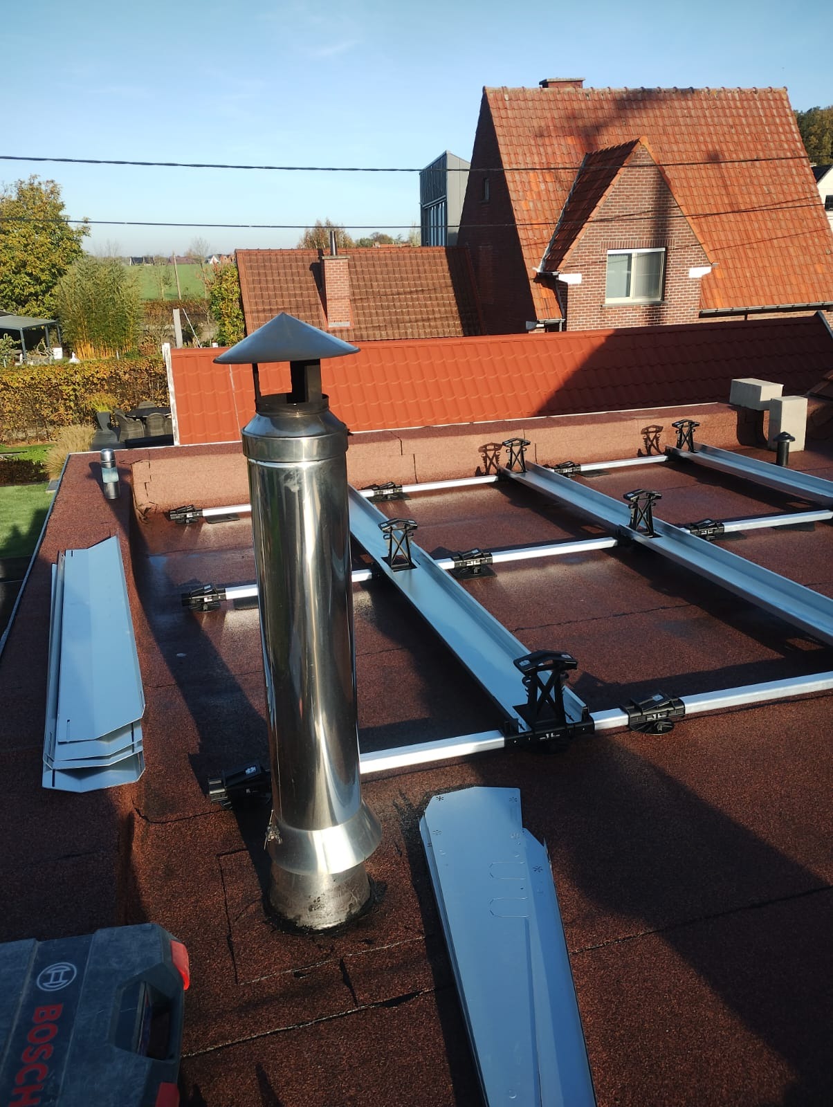 Montaż konstrukcji wsporczej pod panele fotowoltaiczne na dachu pokrytym papą, widoczny komin z nierdzewnej stali i elementy mocujące szyny.