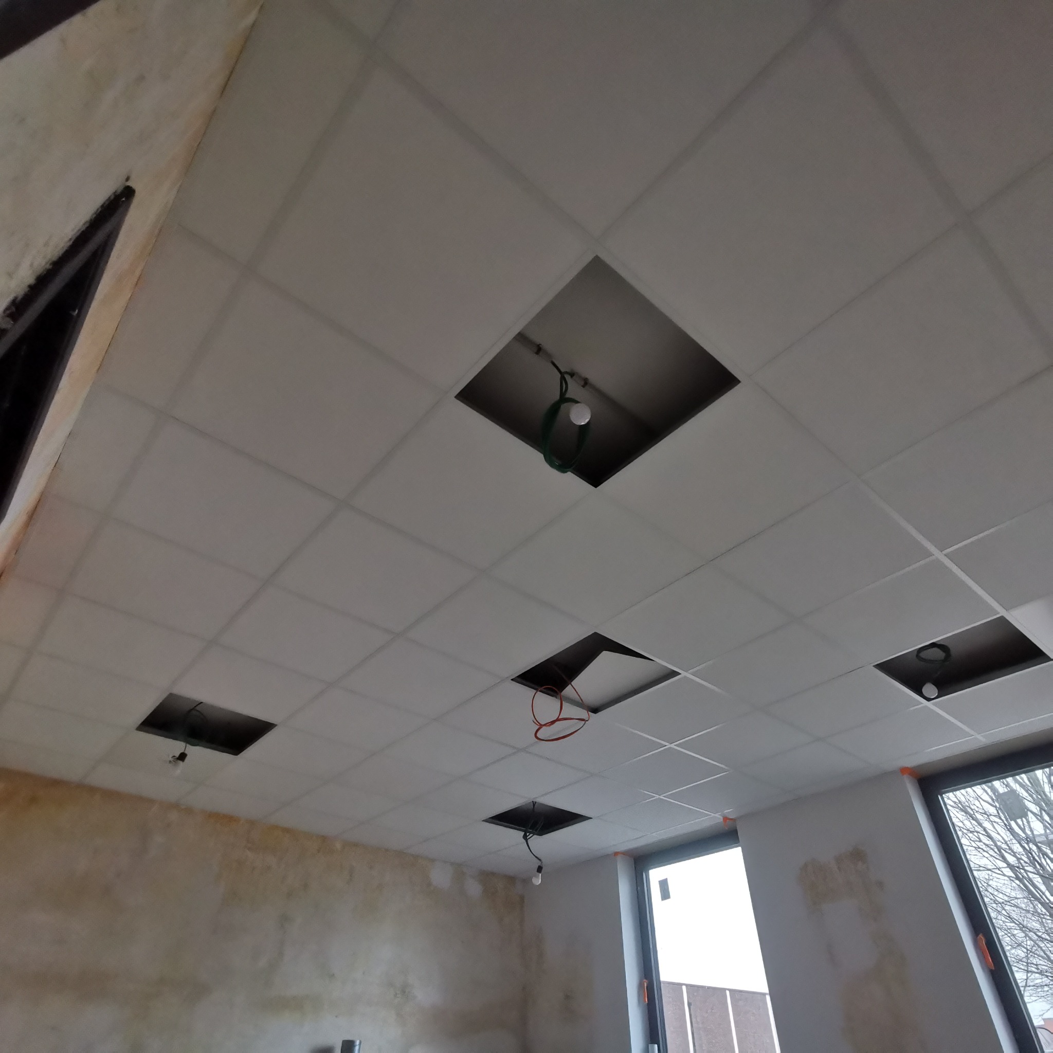 Sufit podwieszany kasetonowy z otworami, widoczne wystające przewody elektryczne i żarówki, ściany w trakcie wykończenia.