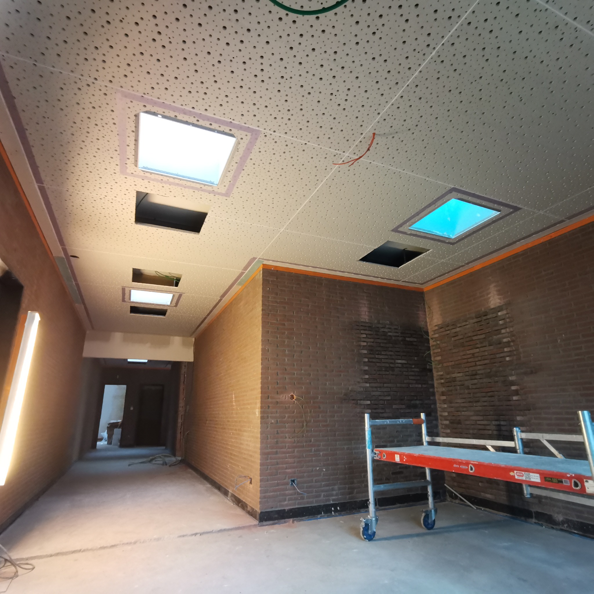 Wnętrze w trakcie remontu: sufit z perforacją i otworami na oświetlenie, fragment instalacji elektrycznej, ceglana ściana, rusztowanie na kółkach.