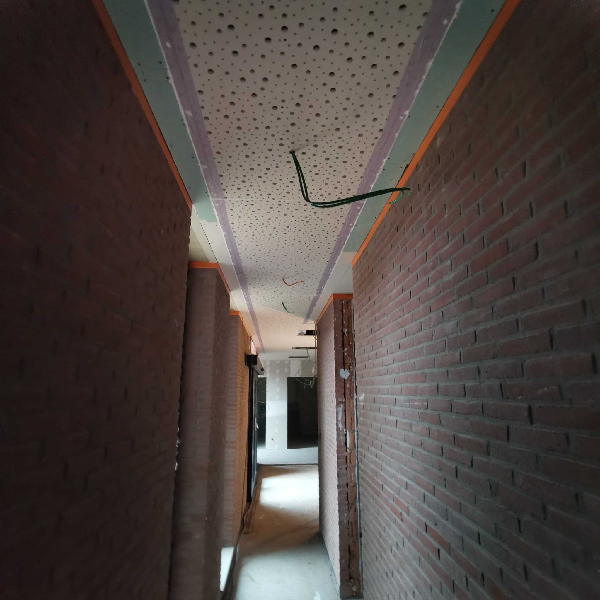 Wnętrze korytarza w trakcie remontu: sufit z perforowanej płyty gipsowej, wystające przewody elektryczne, ściany z cegły, widoczne elementy konstrukcyjne.