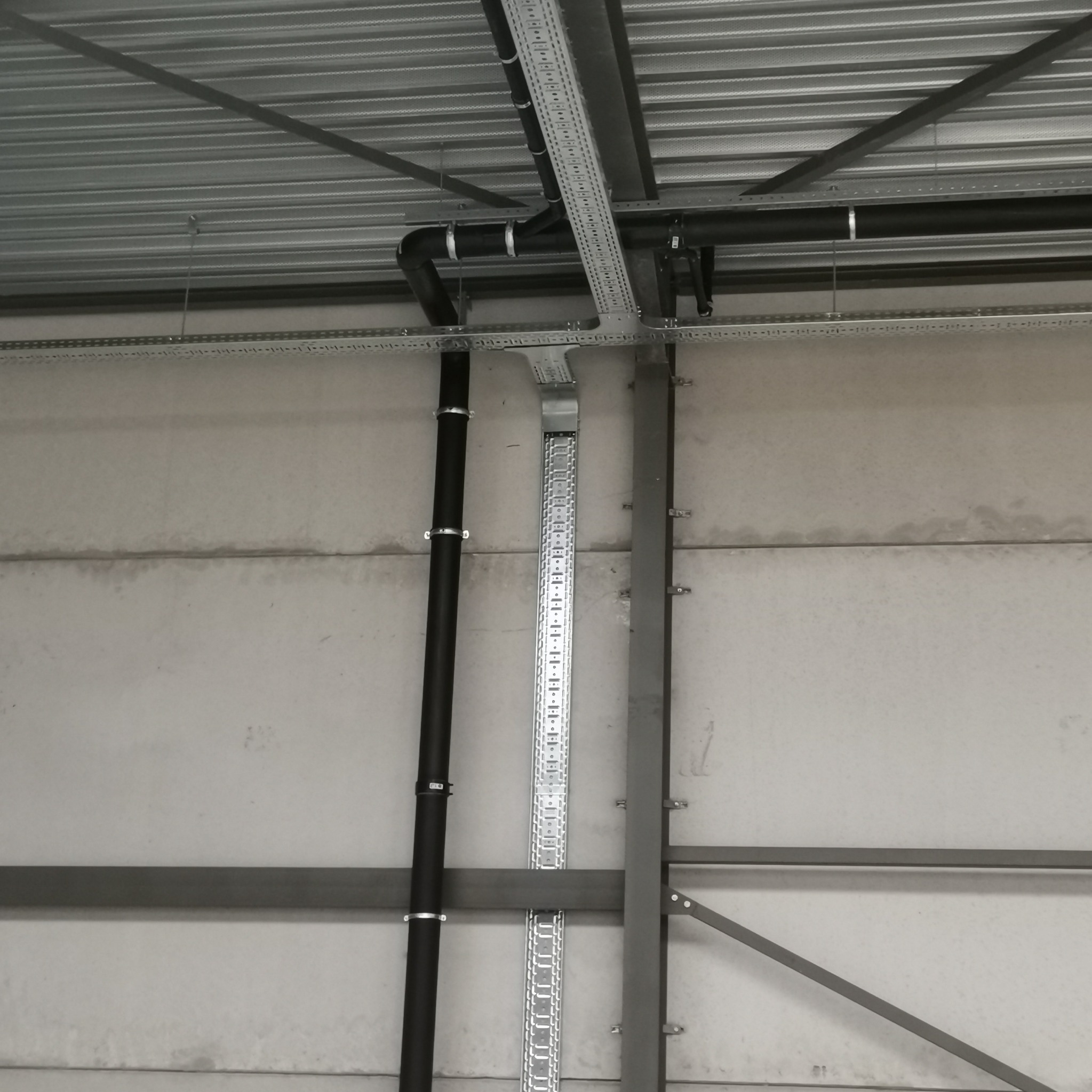 Metalowa konstrukcja sufitowa z zamontowanymi czarnymi rurami i perforowanymi korytkami kablowymi w hali przemysłowej.