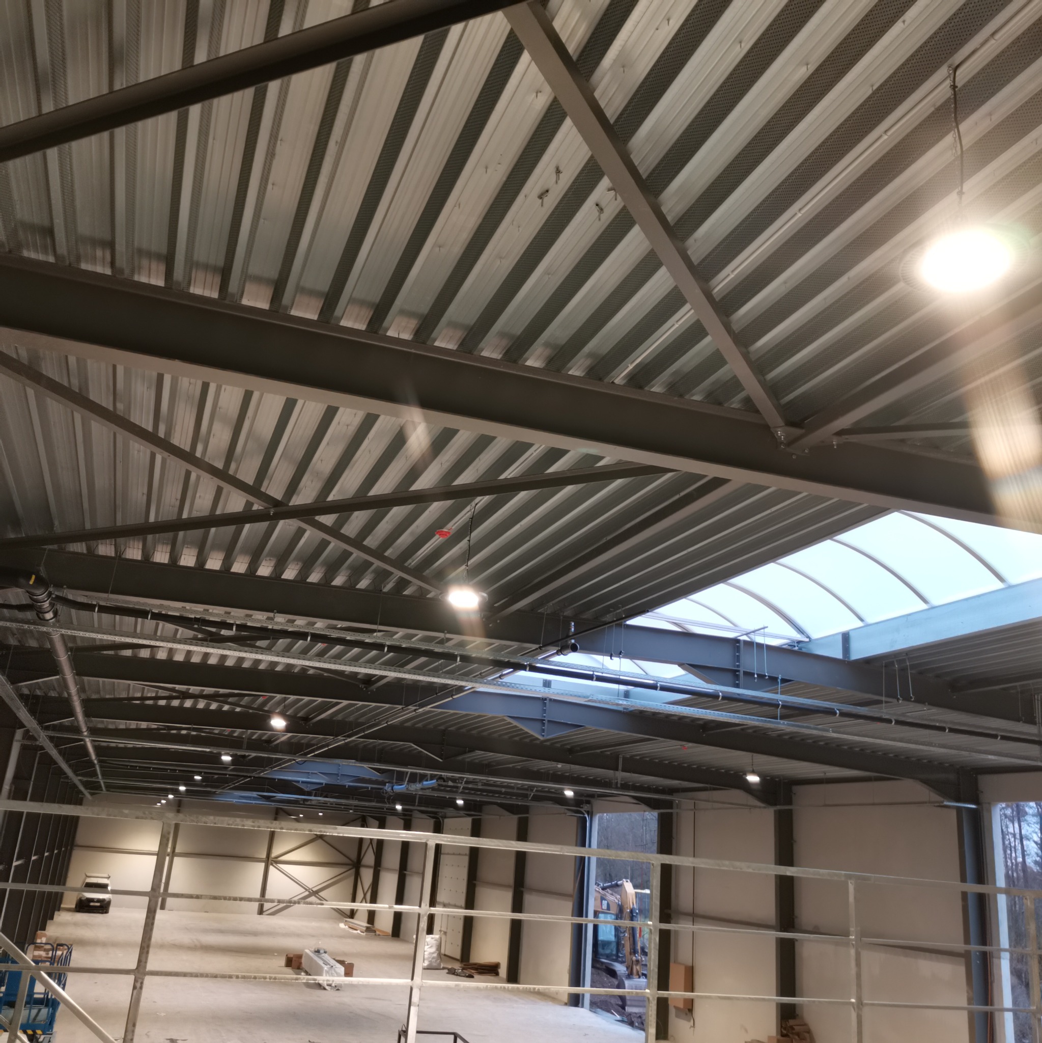 Wnętrze hali produkcyjnej w trakcie budowy z widocznym stropem z blachy trapezowej, stalową konstrukcją nośną, oświetleniem przemysłowym i świetlikiem dachowym.