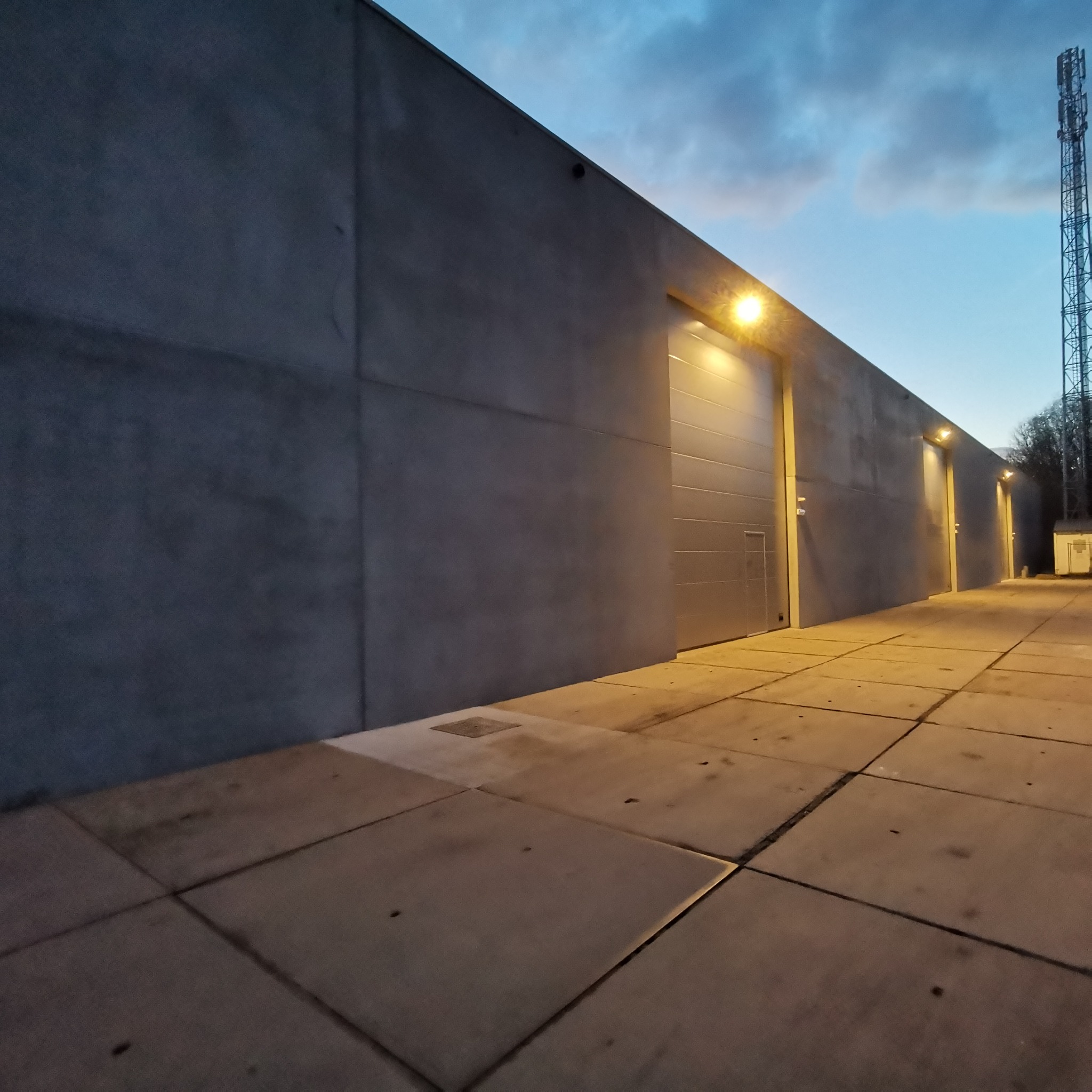 Oświetlenie zewnętrzne budynku przemysłowego z betonową elewacją i segmentowymi bramami garażowymi podświetlonymi lampami, widok z perspektywy chodnika z betonowej kostki.