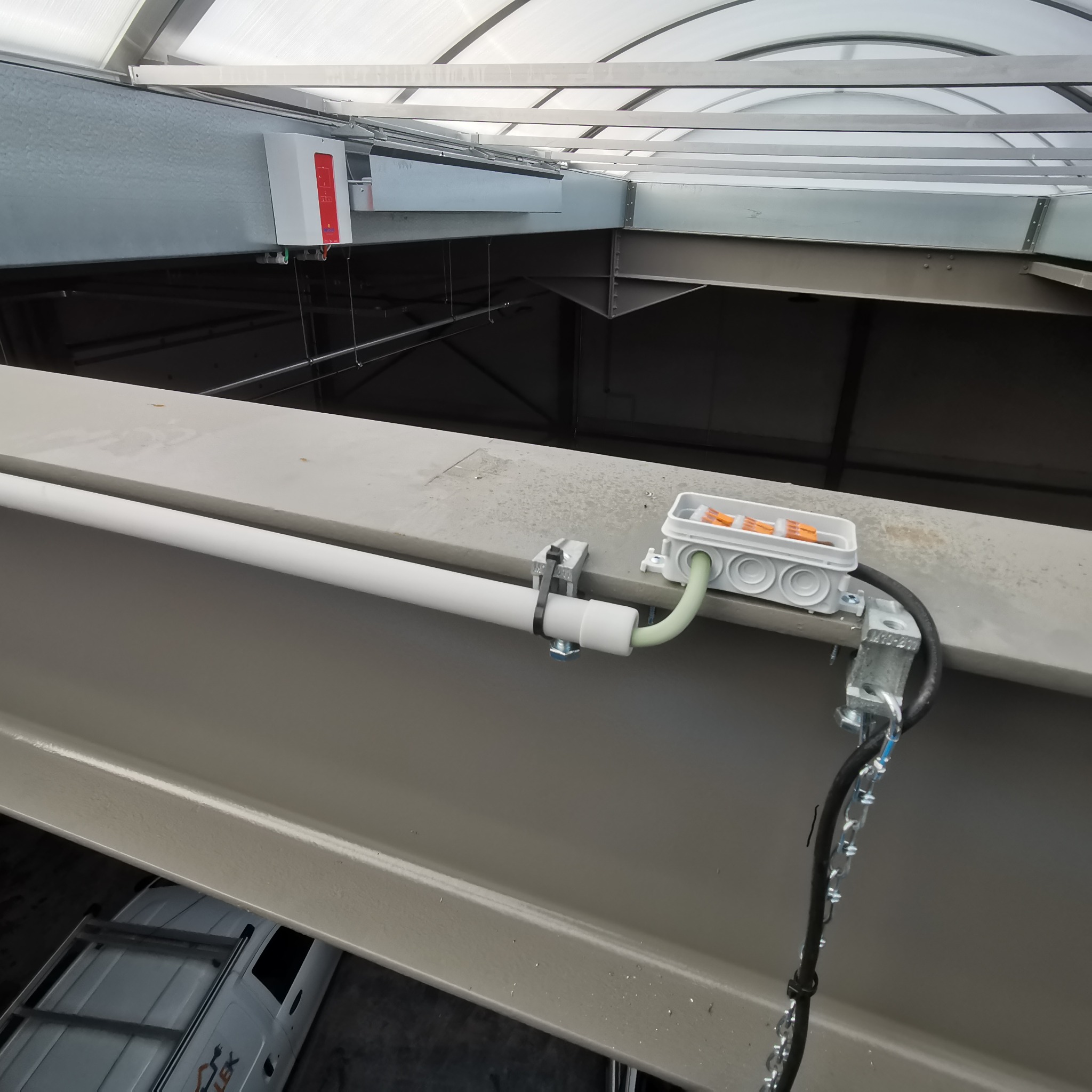 Biała rura przymocowana do metalowej konstrukcji za pomocą uchwytu, z podłączoną szarą puszką elektryczną z pomarańczowymi złączkami wewnątrz, widok z góry na tle konstrukcji dachu hali.