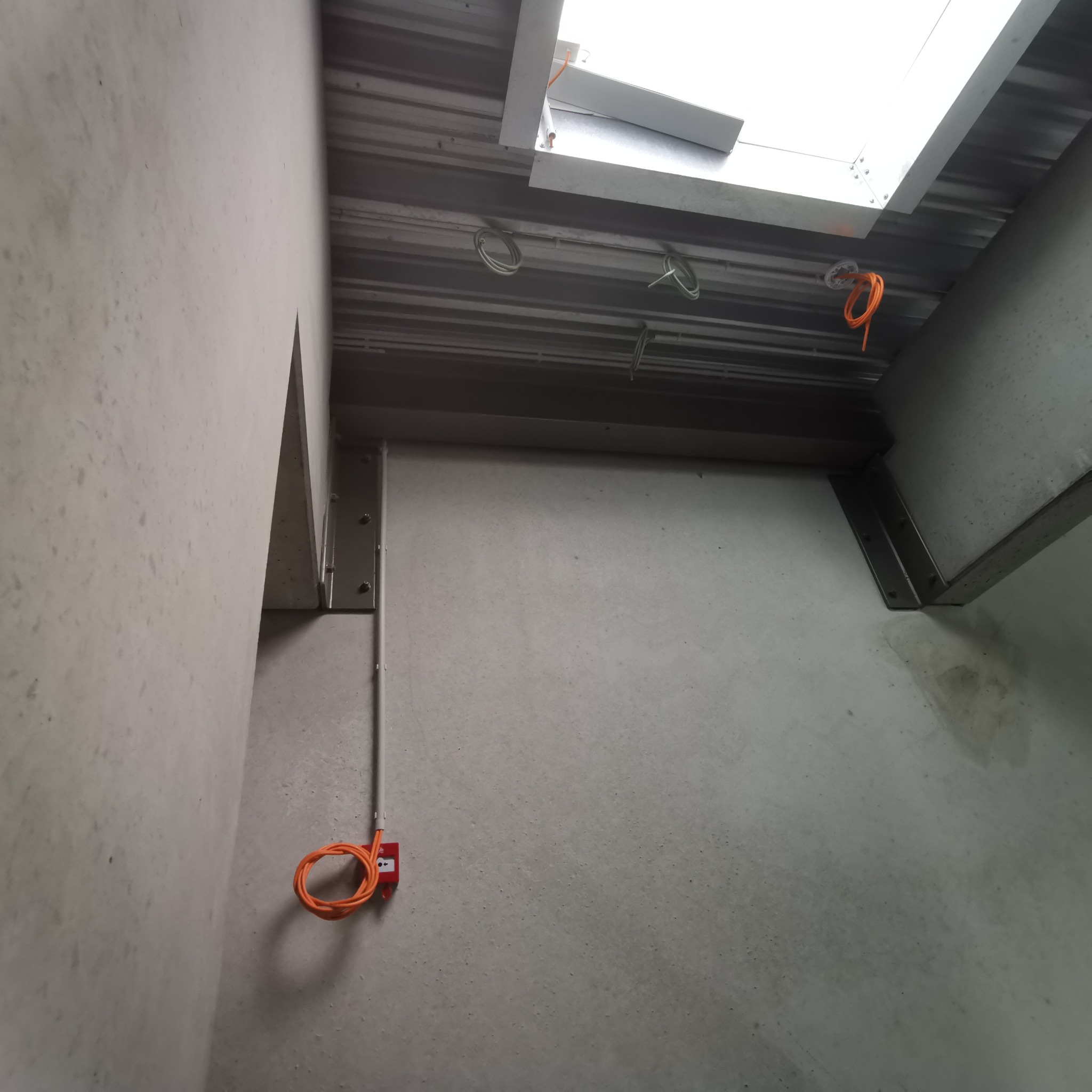 Surowe wnętrze z widoczną instalacją elektryczną: pomarańczowe przewody zwinięte w pętle na ścianie i pod sufitem, szare betonowe ściany i sufit z metalowymi elementami konstrukcyjnymi...