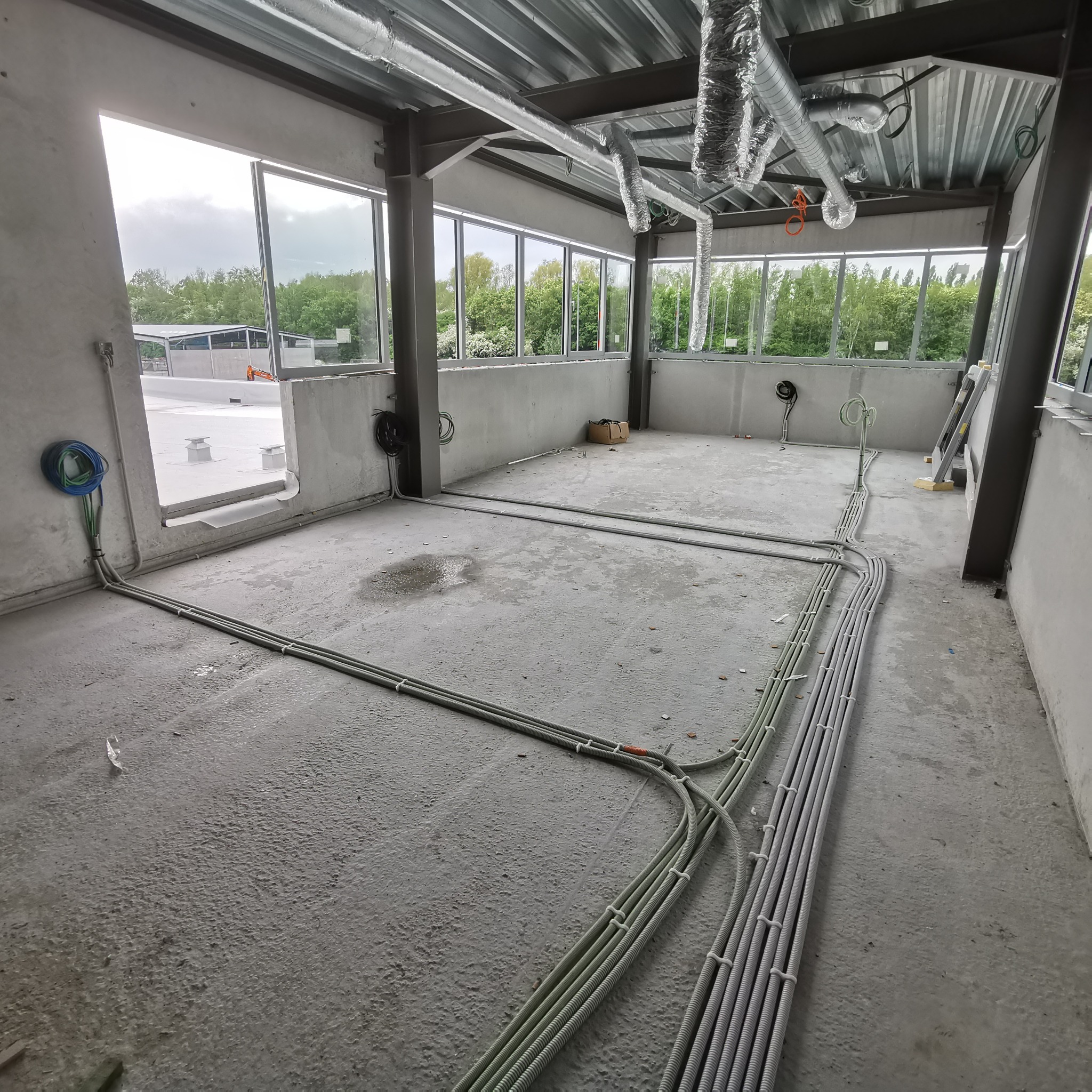 Wewnętrzne okablowanie elektryczne w surowym stanie budynku, widoczne peszle prowadzone po podłodze i ścianach, z oknami w tle i wentylacją pod sufitem.