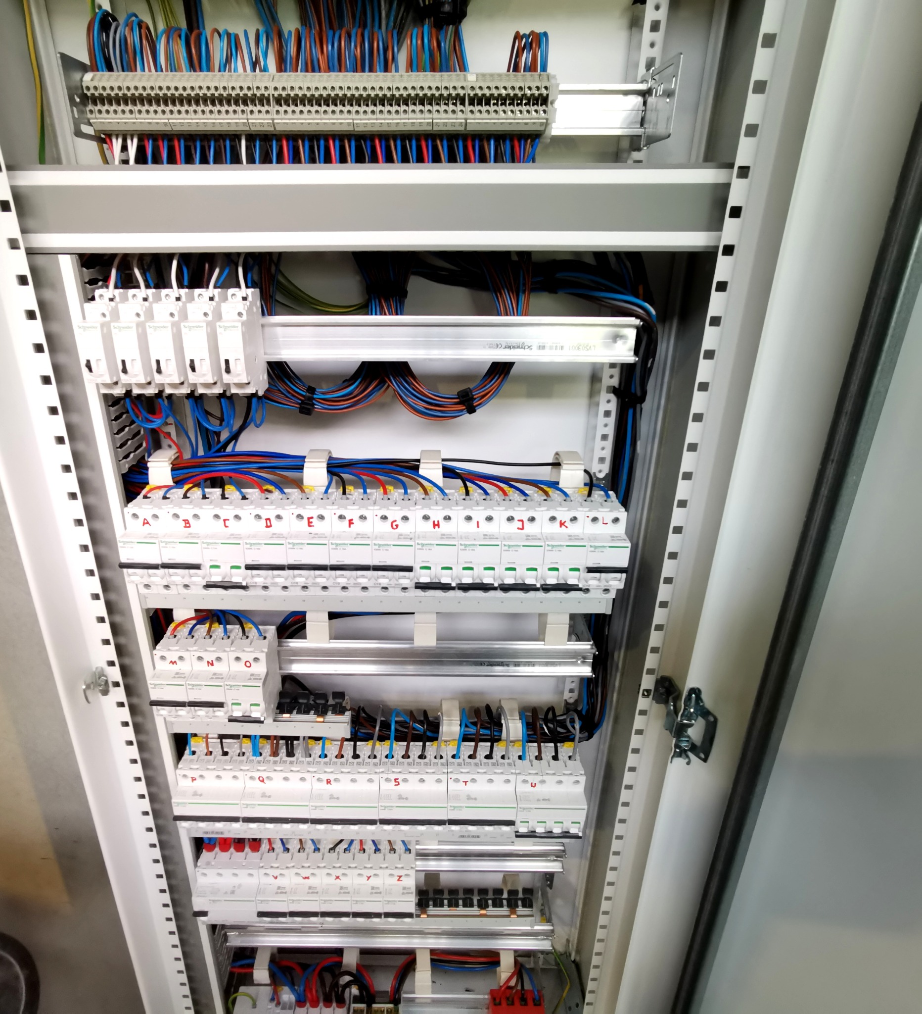 Rozdzielnica elektryczna w metalowej obudowie z widocznymi bezpiecznikami, przewodami oznaczonymi literami od A do Z oraz listwami zaciskowymi.