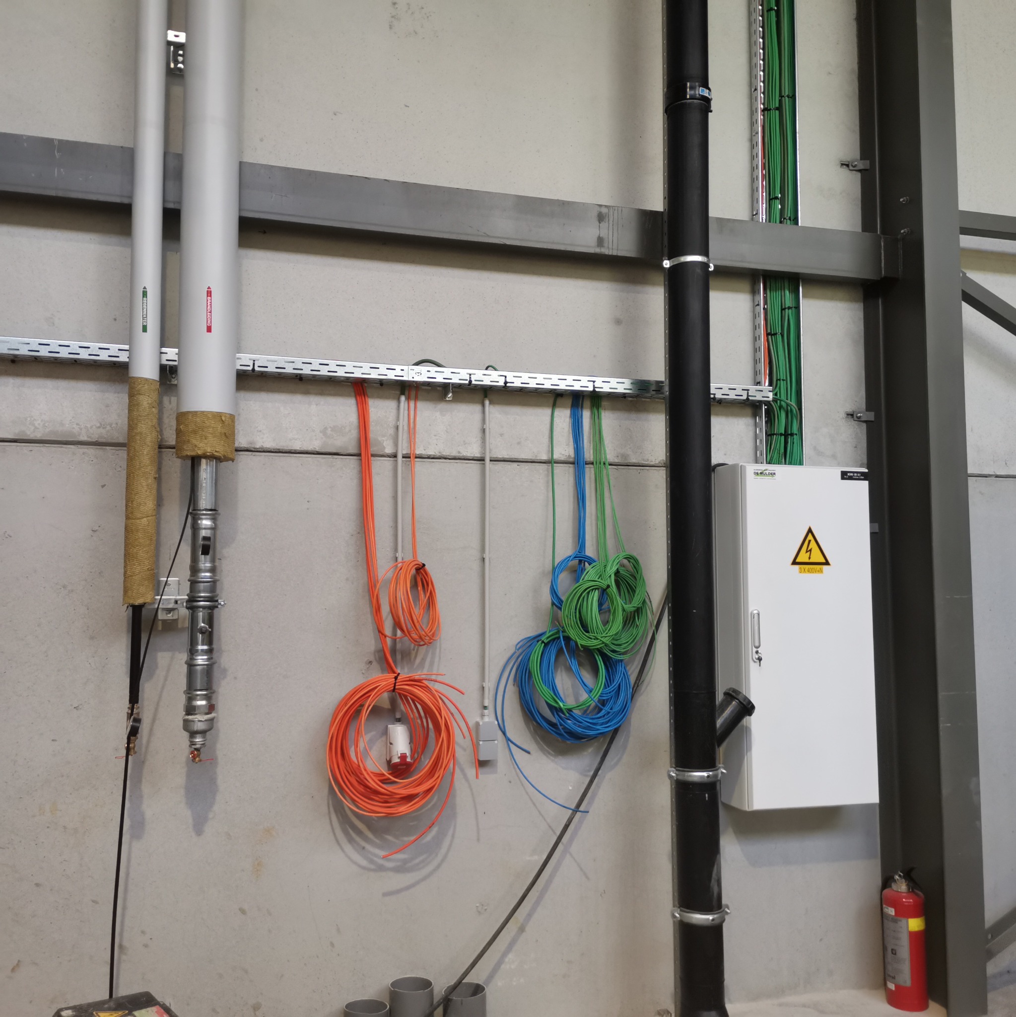 Wnętrze w Kościelcu z widocznymi rurami instalacyjnymi, przewodami elektrycznymi zwisającymi ze ściany na metalowym wsporniku oraz skrzynką elektryczną z symbolem ostrzegawczym.