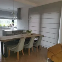 Szara roleta pozioma zamontowana w oknie w nowoczesnym wnętrzu, widoczny drewniany stół z krzesłami i fragment kuchni w tle.