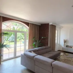 Beżowe zasłony na szynach zamontowane w przestronnym salonie z dużym łukowym oknem wychodzącym na ogród, ceglana ściana w tle.