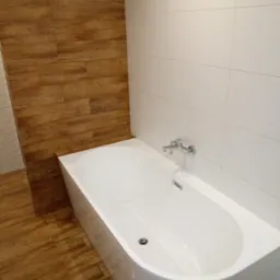 Nowoczesna łazienka z białą wanną, chromowaną baterią i ścianami wyłożonymi białymi płytkami oraz panelami imitującymi drewno.