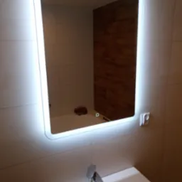 Nowoczesne lustro łazienkowe z podświetleniem LED, odbijające fragment ściany z panelami imitującymi drewno, umywalka z chromowaną baterią