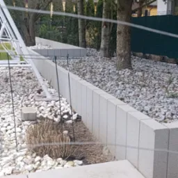 Betonowe murki oporowe tworzące rabaty wypełnione białym i szarym grysem, widoczne drzewa w tle.