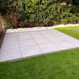 Jasnoszara, kwadratowa powierzchnia z płytek w ogrodzie, otoczona zieloną trawą i bujną roślinnością. Wykończona białym kamieniem dekoracyjnym.