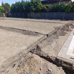 TRANSTECHNIK DZMITRY KARAVAI - Przygotowany teren pod przyszły ogród z wykopanymi rowami, widoczny fragment kostki brukowej i osoba pracująca w oddali, słoneczny dzień.