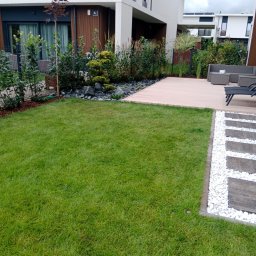 TRANSTECHNIK DZMITRY KARAVAI - Nowoczesny ogród z trawnikiem, drewnianym tarasem i ścieżką z białych kamieni. W tle budynek z drewnianą elewacją i roślinnością ozdobną.