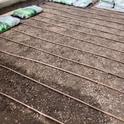 TRANSTECHNIK DZMITRY KARAVAI - Montaż systemu nawadniania w szklarni: brązowe rury ułożone równolegle na ziemi, worki z ziemią w tle. Widoczne złączki i elementy instalacji.