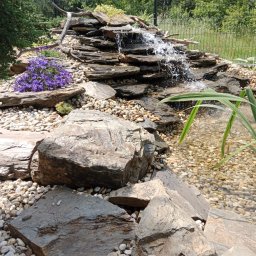 TRANSTECHNIK DZMITRY KARAVAI - Kaskada wodna z kamieni i roślin w ogrodzie. Woda spływa po ułożonych warstwowo płytach skalnych do małego zbiornika z jasnymi kamykami. Fioletowe kwiaty i zielona roślinność.