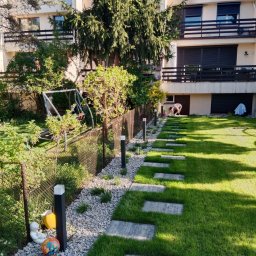 TRANSTECHNIK DZMITRY KARAVAI - Ogród z ścieżką z płyt na trawniku, oświetleniem i ozdobnym żwirem. W tle budynek mieszkalny i osoba pracująca w ogrodzie. Widok na zadbany teren zielony.