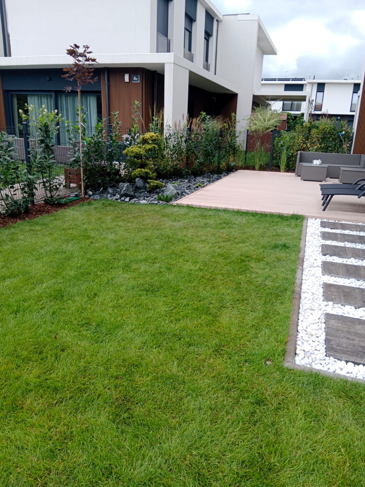 Nowoczesny ogród z trawnikiem, drewnianym tarasem i ścieżką z białych kamieni. W tle budynek z drewnianą elewacją i roślinnością ozdobną.