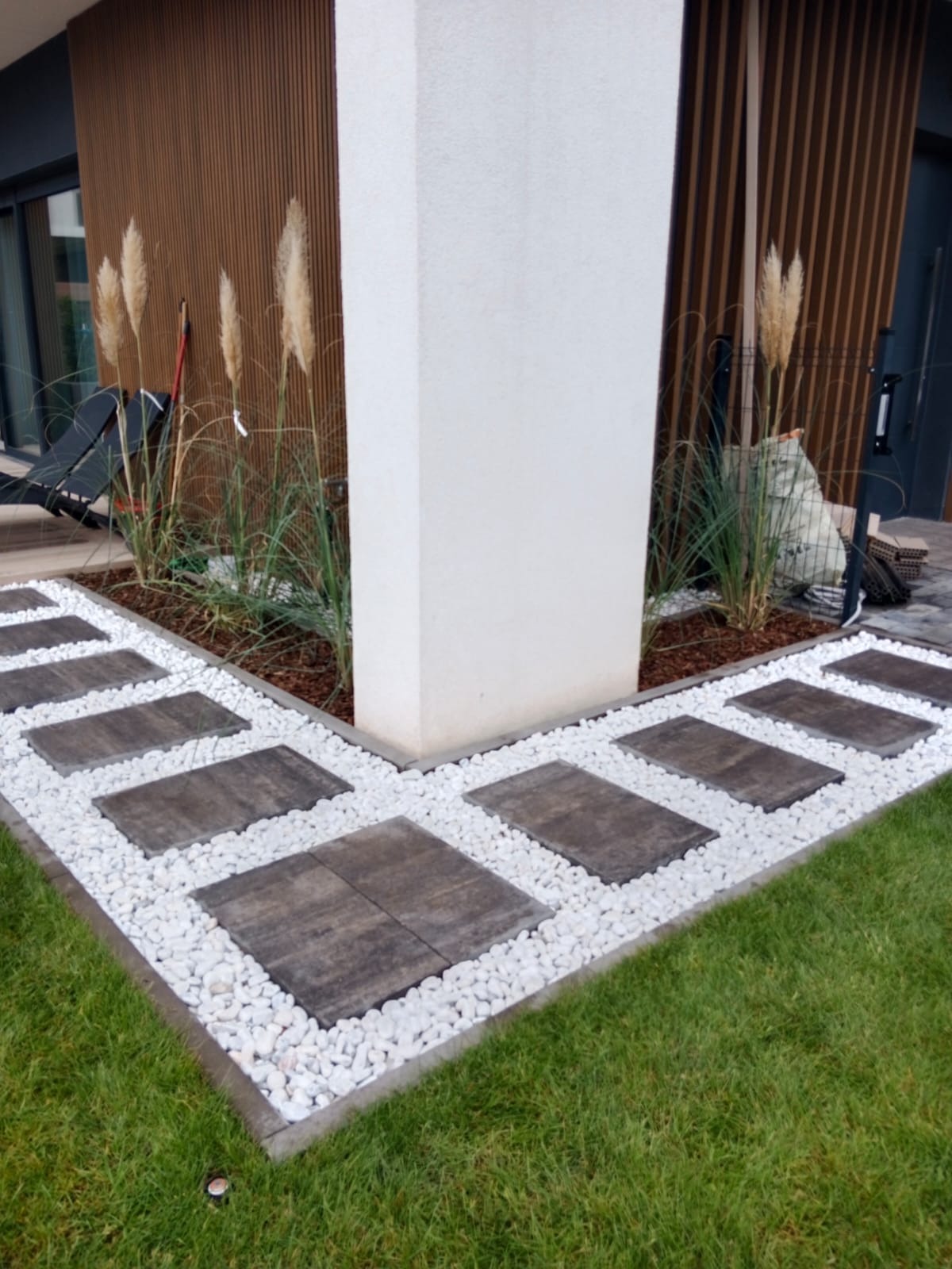 Ścieżka z ciemnych płyt i białych kamieni w ogrodzie, obok budynku z drewnianą elewacją i ozdobnymi trawami. Nowoczesny design ogrodu.