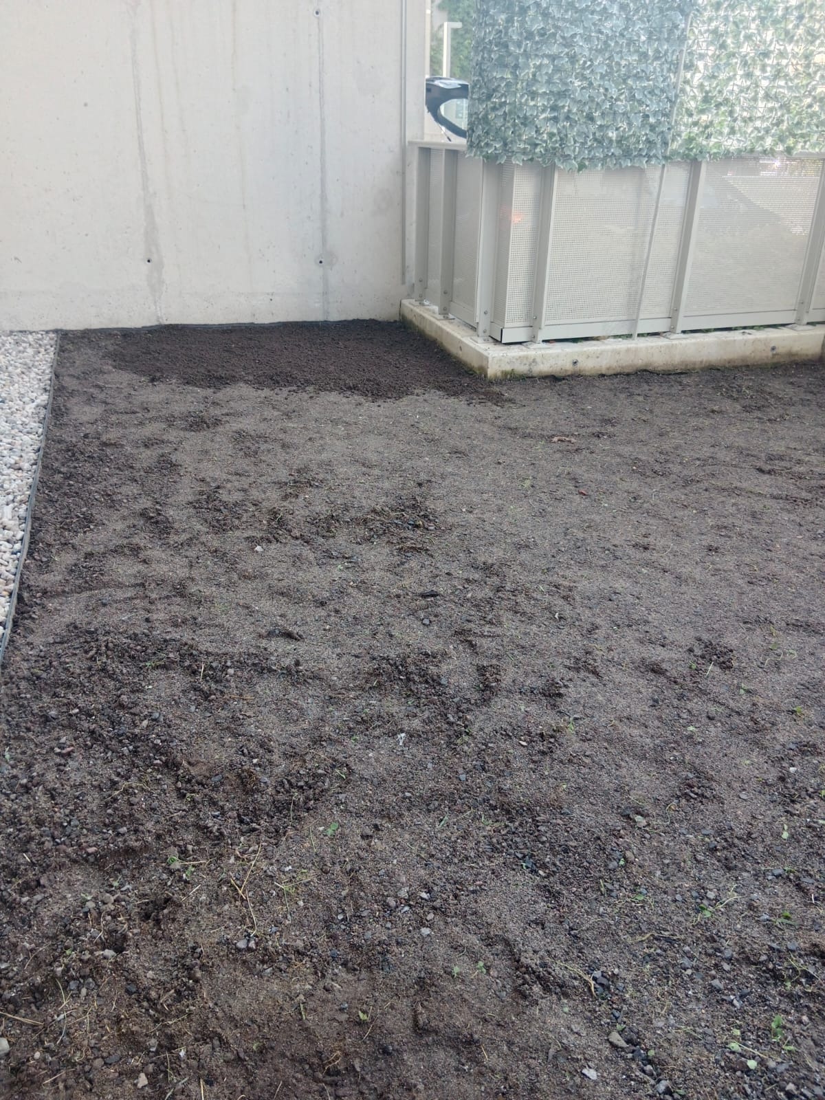 Przygotowany teren pod trawnik z widoczną warstwą świeżej ziemi i obrzeżem z kamieni. W tle betonowa ściana i metalowa konstrukcja z zieloną siatką.