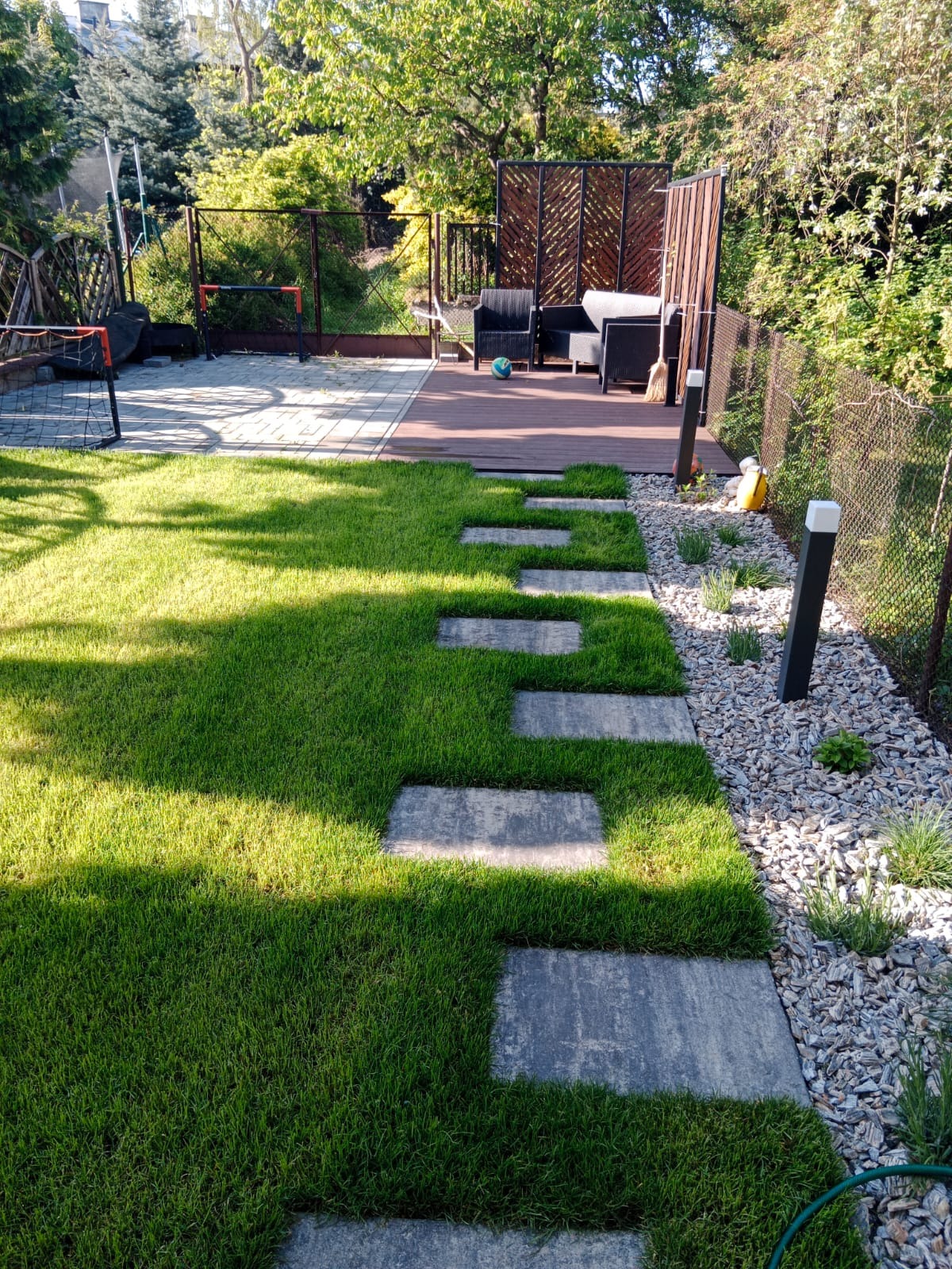 Ścieżka z płyt w trawie prowadząca do drewnianego tarasu z meblami ogrodowymi. Obok trawnika pas żwiru z lampami. W tle siatka do piłki nożnej i zieleń.