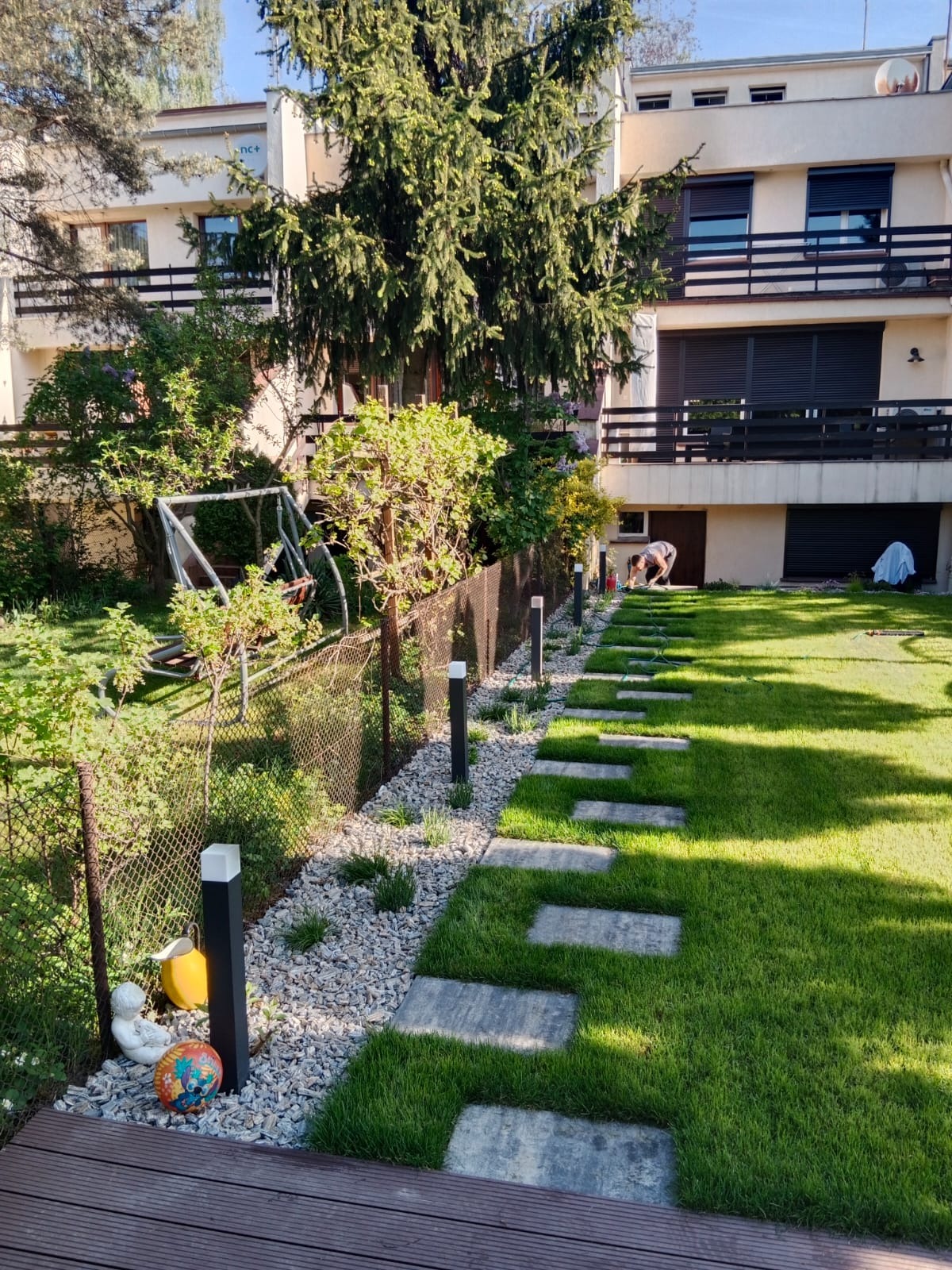 Ogród z ścieżką z płyt na trawniku, oświetleniem i ozdobnym żwirem. W tle budynek mieszkalny i osoba pracująca w ogrodzie. Widok na zadbany teren zielony.